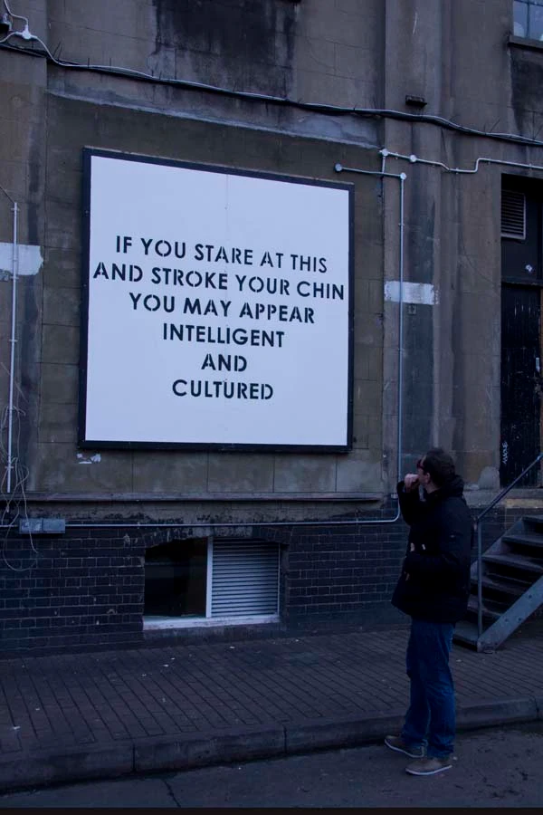 A man is standing in front of a billboard, staring at it and stroking his chin. The billboard reads in block stencil letters "If you stare at this and stroke your chin you may appear intelligent and cultured" 