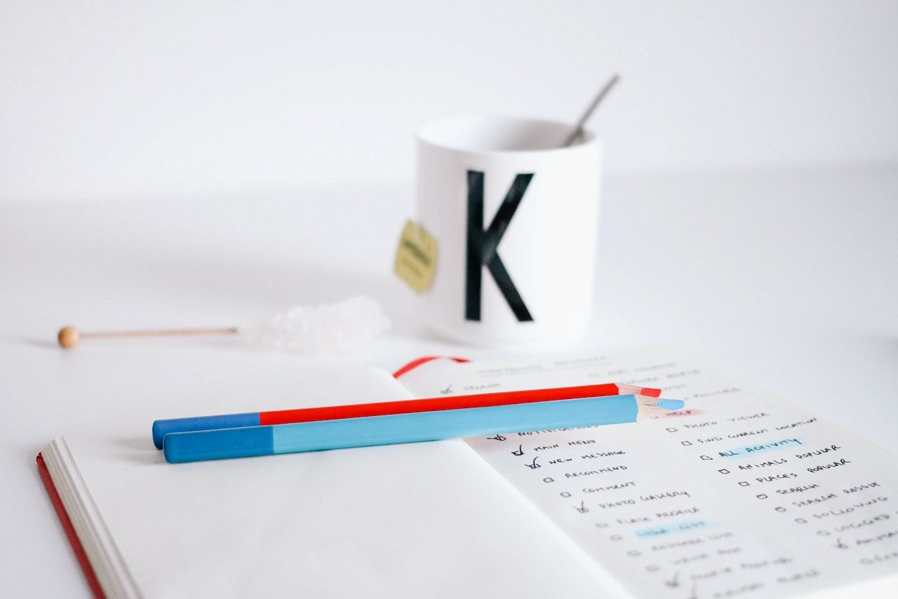 A notebook with a tasklist and two pencils, red and blue in the foreground. In the background there's a tea mug with a letter K and further out a white wall. 