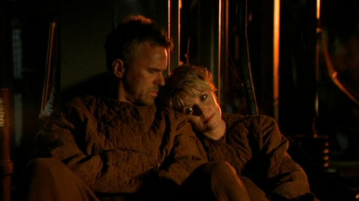 Screencap from the episode showing O'Neill and Carter in their drab worker clothes, sitting on the ground against a backdrop of dimly lit exposed metal pipes and ducting. They look positively despondent. Carter is leaning on O'Neill and resting her head on his shoulder. 