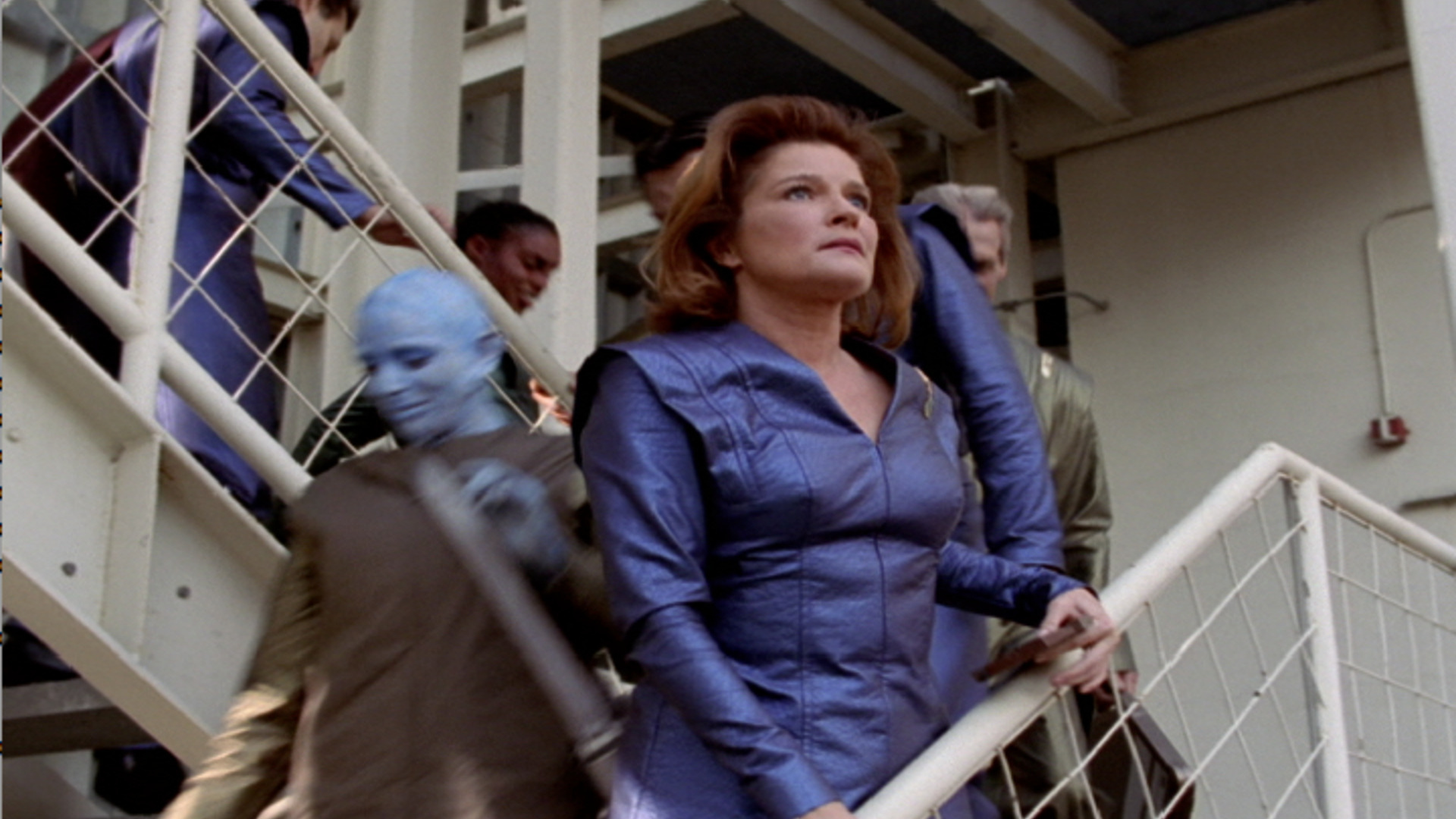 Captain Janeway with her memories erased arrives for her first day at work. She's wearing a blue uniform and standing on an industrial-looking stairway holding the bannister and looking out.